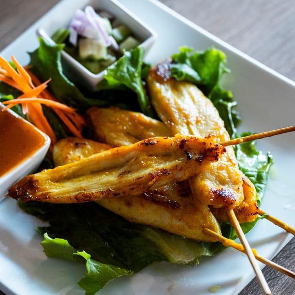 Grilled chicken satay skewers with peanut sauce, cucumber relish, and fresh lettuce on a white plate.